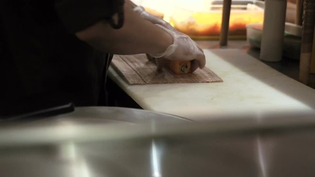 Process of making and cutting orange sushi rolls. Man rolling up sushi set using bamboo mat. Prepared sushi rolls pass through on foreground