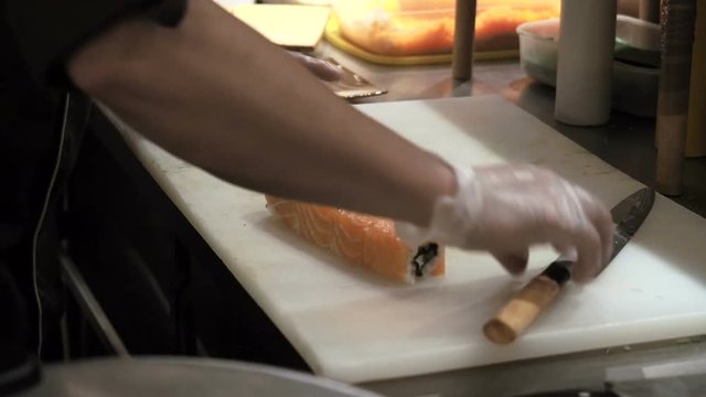 Process of cutting orange sushi rolls by knife. Man rolling up sushi set using bamboo mat. Prepared sushi rolls pass through on foreground