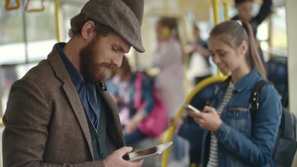 Young man with beard standing in trolleybus and texting someone on tablet, woman communicating on phone in background - Powered by Adobe