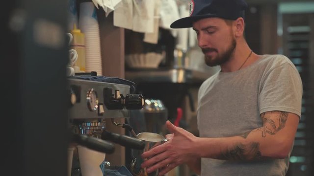 Making coffee. Barista heats milk. Frothing of milk. Coffee Machine. Barista-hipster. A cozy coffee shop. Barista - a young man with tattoos. The tattoo on his arm.