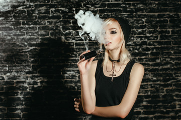 Sexy goth girl smokes electronic cigarette on dark background. The model vaper vaping a vaporizer...