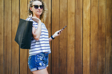 Girl with blond curly hair, dressed in a striped T-shirt and denim shorts, standing on the street on a background of a wooden wall, with his shopping bag over her shoulder while holding a smartphone.