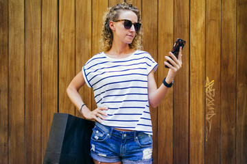 Girl with blond curly hair,dressed in T-shirt and shorts,standing in street and looking at screen of smartphone,is in her hand.In other hand black shopping bag. In background wooden wall. Shopping.