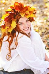 Naklejka na ściany i meble Beautiful young girl in a wreath from yellow leaves, in white clothes, sitting on fall leaves on a Sunny day and laughing. Portrait. Close-up. Vintage processing. Vertical view