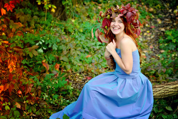 Obraz na płótnie Canvas Charming girl in a wreath from autumn leaves, in a blue dress sitting on a log on the background of red and green bushes in a Sunny day and cheerfully laughs. Portrait. Horizontal view