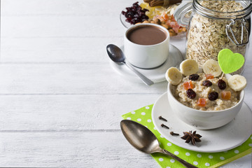 Oatmeal porridge with dried fruits, cranberries, bananas and spices. Cup of cacao and jar of oatmeal on background. Arranged on white background.
