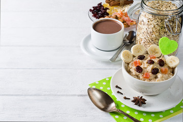 Oatmeal porridge with dried fruits, cranberries, bananas and spices. Cup of cacao and jar of oatmeal on background. Arranged on white background.