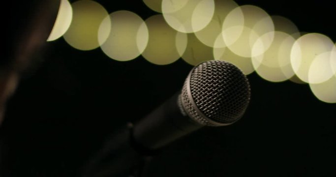 Microphone on a stage with bright lights in the background. Woman with studio headset walking by and knocking at the mic with her finger to check if it works. Slow motion.