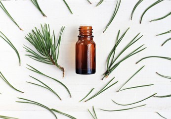 Bottle of essential oil, pine tree twigs and green needles scattered framed, top view, white wooden surface background. 