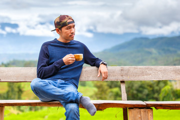 Asian young man smiling with hat sitting on a wooden bench and holding coffee cup in morning time at green terraced rice field in pa pong pieng , Mae Chaem, Chiang Mai, Thailand