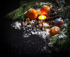 Fototapeta na wymiar Pine branches, candle, Christmas tree decorations, tangerines, nuts, the pine cone in snow on a black background