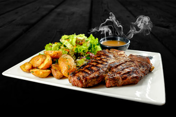 Plate with thick juicy stake, bowl of red sauce, golden french fries and green vegetables. Studio shot with black wooden background.