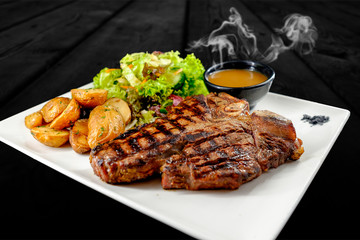 Plate with thick juicy stake, bowl of red sauce, golden french fries and green vegetables. Studio shot with black wooden background.