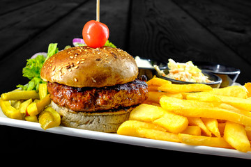 Plate with beef burger, bowl of red, white sauce, golden french fries, pickles and green vegetables. Studio shot with black wooden background.