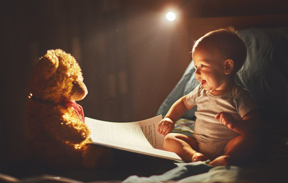 Happy baby reading a book with teddy bear in bed