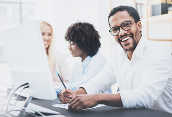 Obraz premium Dark skinned entrepreneur wearing glasses, working in modern office.African american man in white shirt smiling at the camera.Horizontal,blurred background