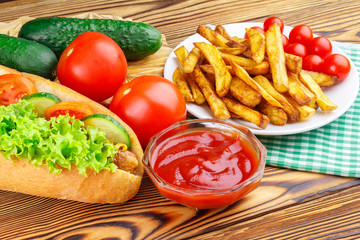 Fast food meal, hot dog, french fries, ketchup, tomato and cucumber on wooden background.