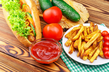 Fast food meal, hot dog, french fries, ketchup, tomato and cucumber on wooden background.