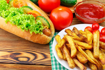 Fast food meal, hot dog, french fries, ketchup, tomato and cucumber on wooden background.