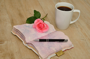 An open diary on a wooden table with a pink rose, pen and a mug