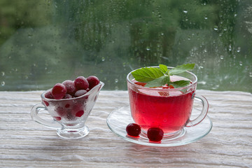 On the street it is raining. Drops on the window glass. On the table is a glass vase with cherries and a Cup of cold drink red. Frost on fruits in the vase. Healthy eating
