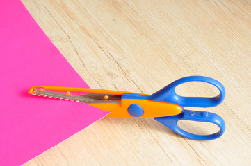 A colorful scissor that cut a zigzag pattern with a pink paper 