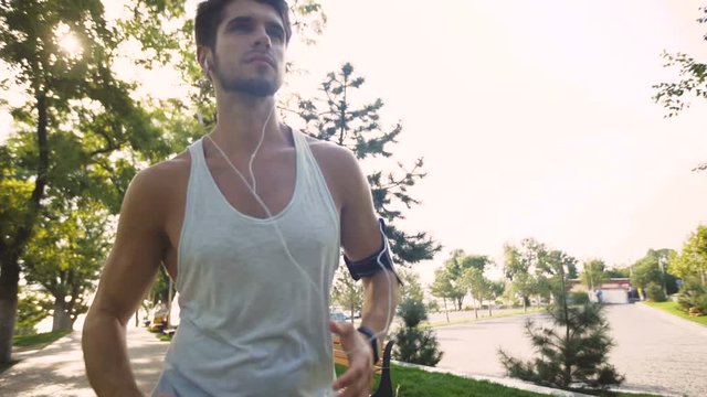 Great active young man jogging on park, while listening to music in earphones from mp3 player smartphone.