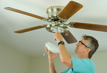 Professional or DIY do-it-yourself home owner doing ceiling fan repair work with the glass cover removed as he adjusts the fixture.