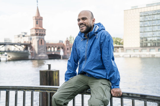 Germany, Berlin, laughing man wearing blue rain jacket sitting on railing in front of Oberbaum Bridge