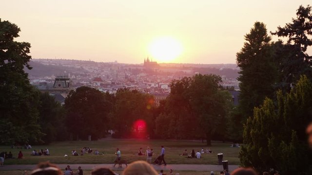 A moving shot behind a crowd of tourists hanging out in Riegrovy Sady park with a view of Prague castle during sunset.  A man walks his dog in the distance. 4k. Slow motion.