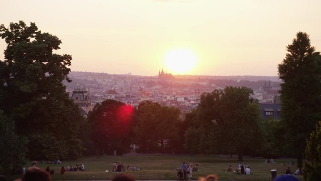 A moving shot behind a crowd of tourists hanging out in Riegrovy Sady park with a view of Prague castle during sunset.  Smoke hang sin the air. 4k. Slow motion.