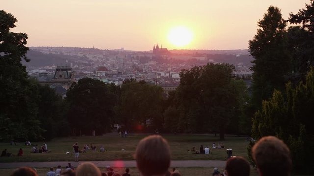 A moving shot behind a crowd of tourists hanging out in Riegrovy Sady park with a view of Prague castle during sunset.  4k. Slow motion.