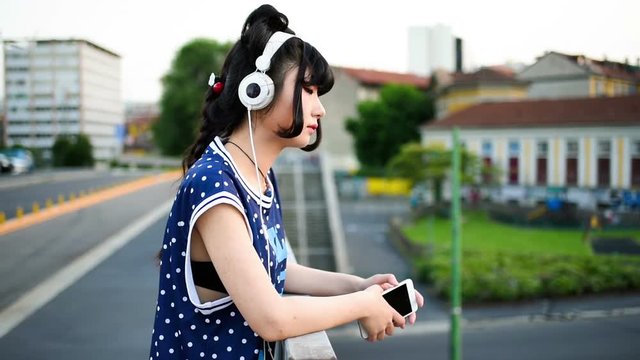 young beautiful asian millennial woman listening music with earphones and smart phone hand hold outdoor in city back light - relaxing, music, technology concept