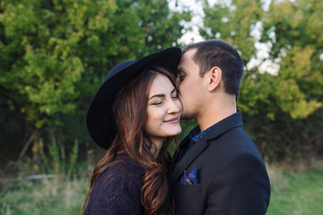 young man in a black suit kisses a young woman in black hat and dress in the park. loving couple in black clothes outdoor. Elegant costumes for Halloween, concept