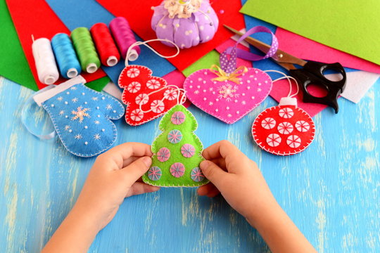 Small child holds felt Christmas tree in his hands. Child shows Christmas crafts. Felt fur-tree, mitten, heart, ball decor on wood background. Flat felt sheets, scissors, sewing set, pins, pincushion