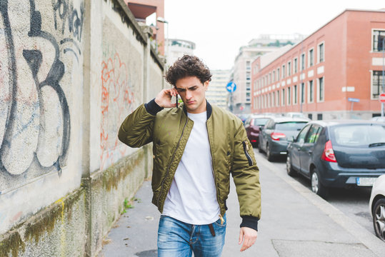 Young handsome cauciasain curly hair man walking in the street of the city, talking smartphone, overlooking pensive - communicaiton, technology, thoughtful concept