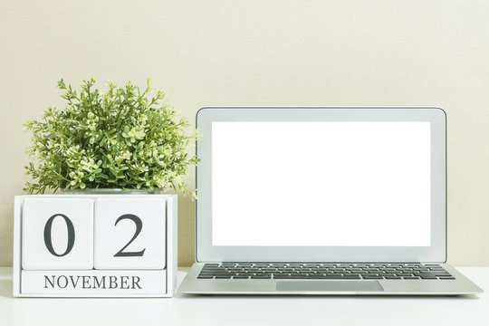 White wooden calendar with black 2 november word with white blank space at center of computer notebook on white wood desk and cream wallpaper textured background , selective focus at the calendar