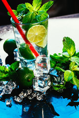 glass of mojito with lime and mint ice cube close-up red straw on blue background