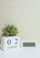 White wooden calendar with black 2 november word with clock and plant on white wood desk and cream wallpaper textured background , selective focus at the calendar
