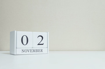 Closeup white wooden calendar with black 2 november word on blurred white wood desk and cream color wallpaper in room textured background with copy space , selective focus at the calendar