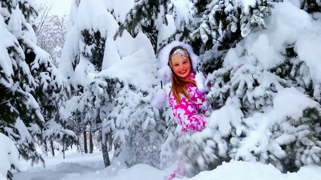 Little kid girl in winter clothes with falling snow near tree. Child plying in winter forest.