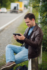 Fototapeta premium Man orders taxi from his tablet sitting on a road fence