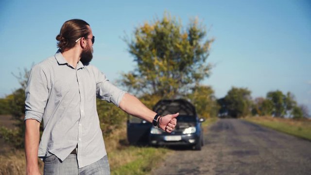 Attractive bearded man thumbing a lift on a rural road with his broken down car pulled onto the verge behind him