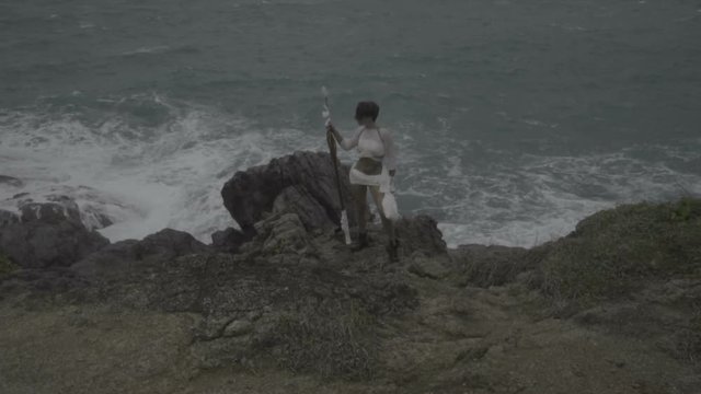 Beautiful amazon woman warrior wearing white outfit standing on the rocks with rifle over stormy sea background - video in slow motion