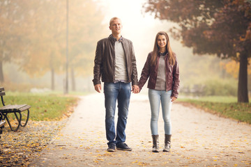 Young couple are walking in a park on a one Autumn day. They are holding heands and smiling and they are looking to camera