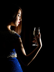 Young celebrating woman in a blue dress holding a glass of champagne on a dark background. play of light and shedow