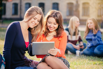 Technology, education, friendship and people concept - two smiling young woman with tablet pc computers sitting on a grass