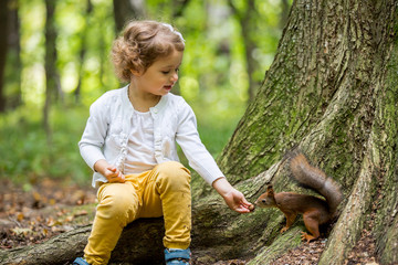 Little beautiful girl sitting on green lawn and feeding squirrel with nuts. Sweet, happy child on a...