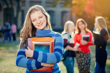 Enjoying university life. Pretty young woman with long hair holding books and smiling while standing against university with her friends chatting in the background out of focus