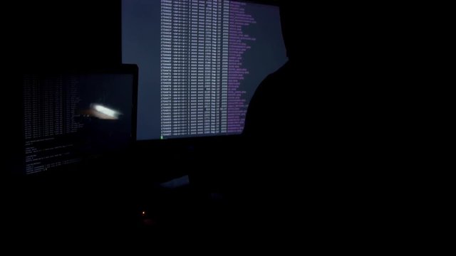 Hacker in hood cracking code using computers in dark room